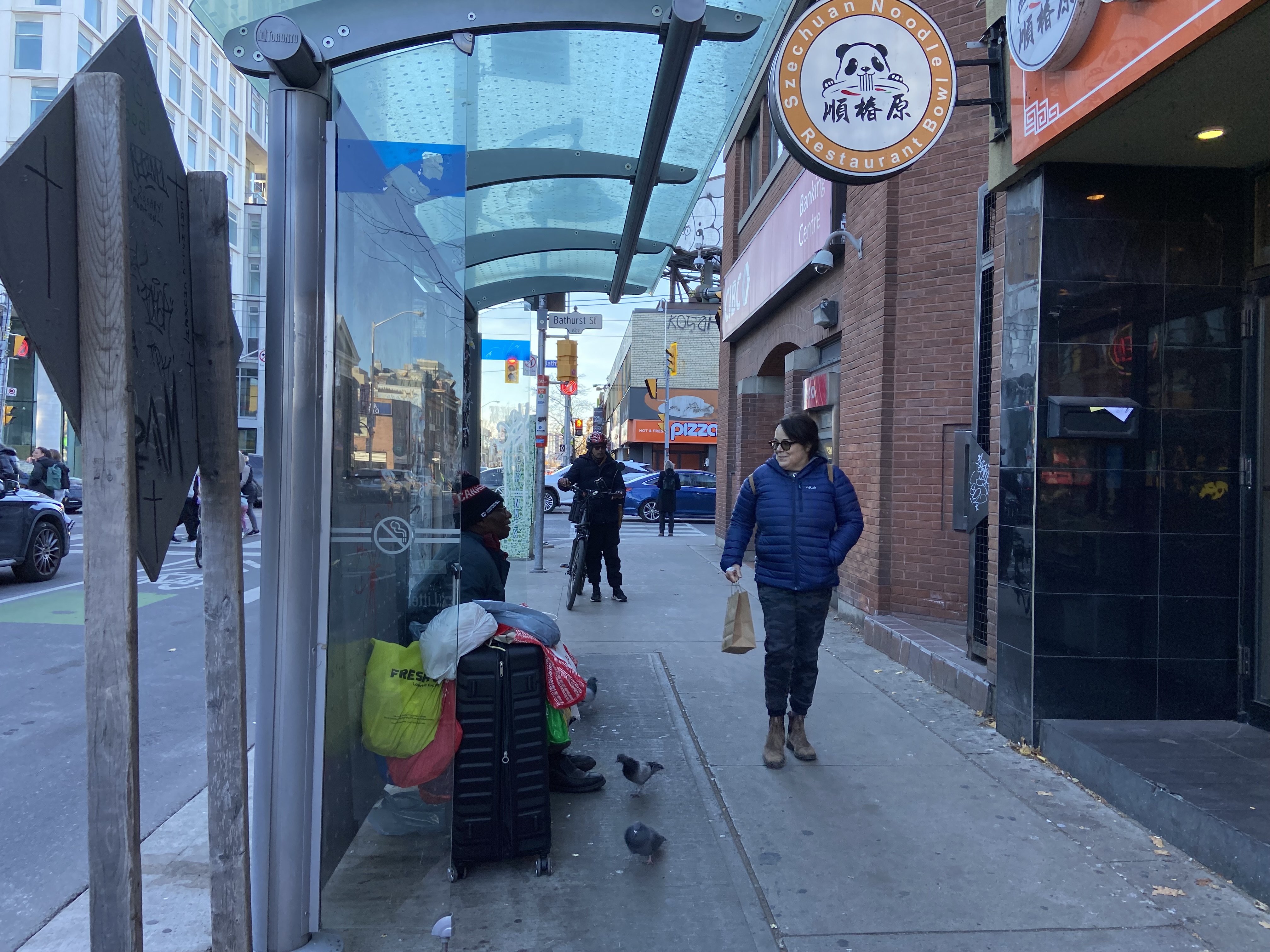 A man sits at a bus stop as a someone passes by on the sidewalk