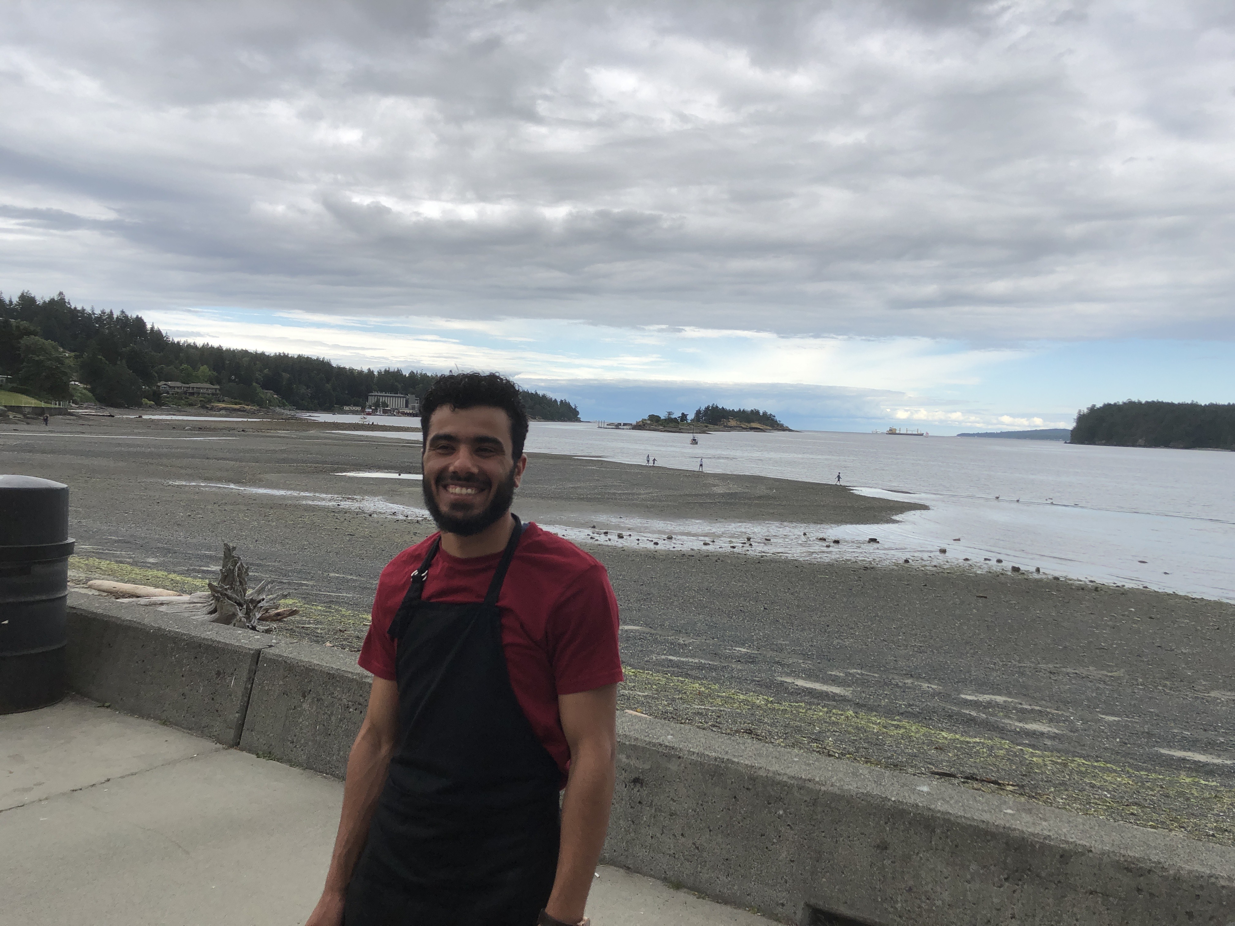 A man with a red shirt and black apron in front of beach and ocean